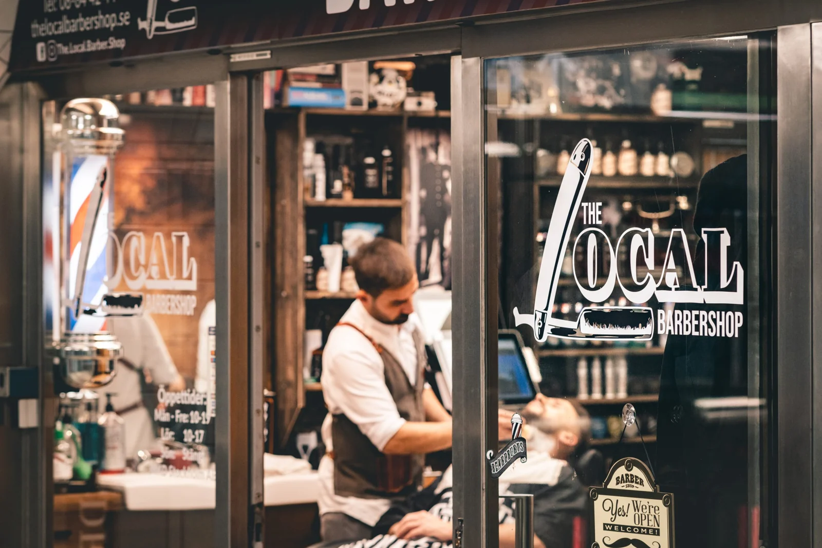 The Local Barbershops skyltfönster med en barberare i arbete innanför